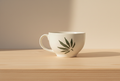 The Verdé Vogue Cup, a luxury ceramic cup with a green cannabis botanical leaf design, rests on a light wooden surface in gentle natural light.